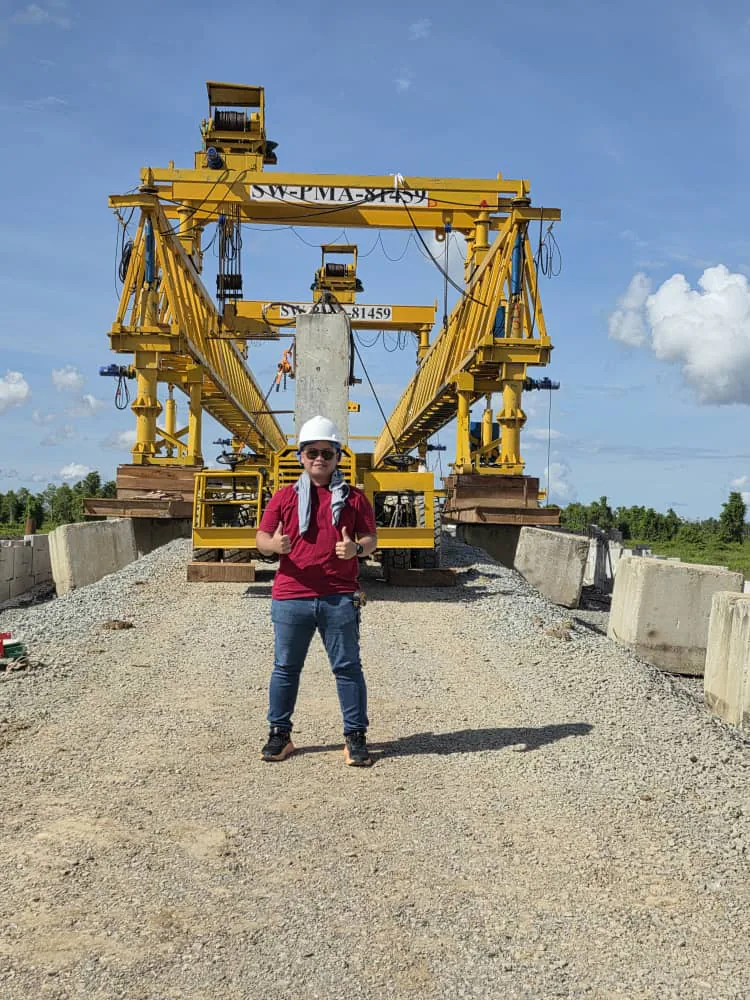 Ir. Denth Ringam on site in a white hard hat, standing on the deck of a bridge under construction in front of a yellow segment-launching gantry.